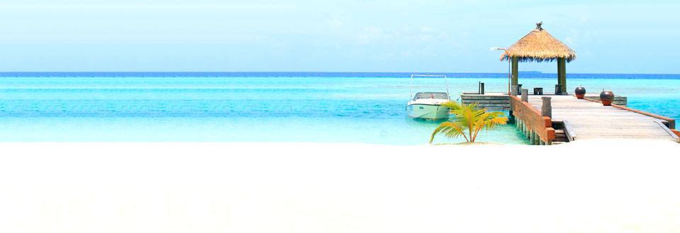 En paradisisk strandvy med vit sand, en träbrygga som leder till en hydda med halmstak och en båt.