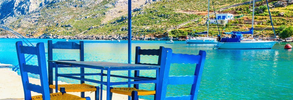 En pittoresk strandvy med blå stolar och bord framför en klarblå vik med båtar och berg i bakgrunden.