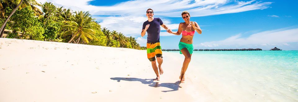 En man och kvinna ler och springer längs en solig strand med palmer i bakgrunden.
