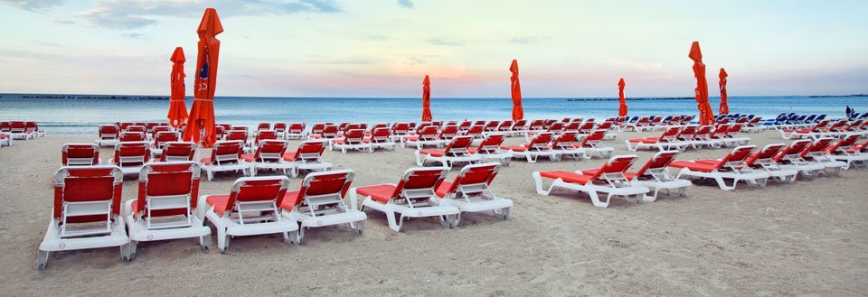 Tomma röda solstolar på en sandstrand med ihopfällda parasoller vid havet i skymningen.