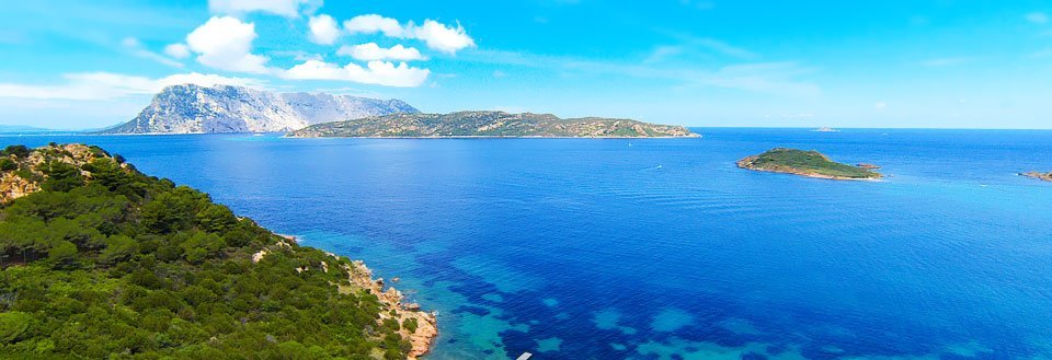 Panoramavy över ett kristallklart blått hav med gröna öar och en bergskedja i bakgrunden under en blå himmel.