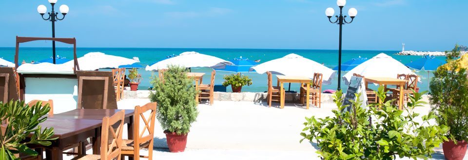 En fridfull strandkrog med trämöbler och vita parasoller framför det glittrande havet.