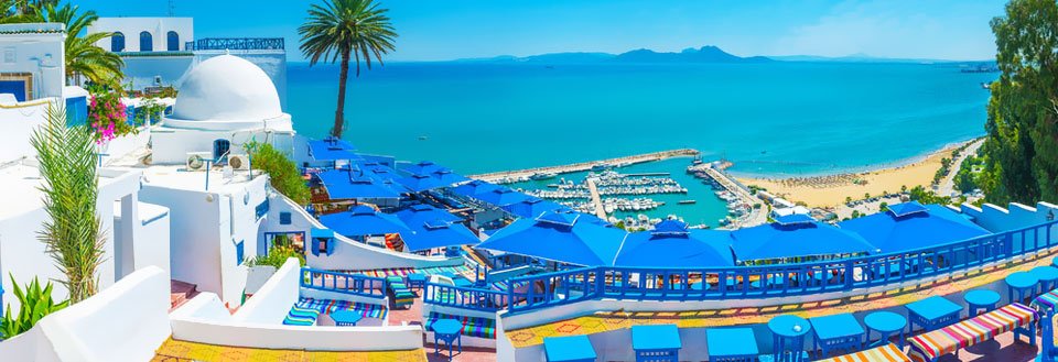 Panoramavy över en solig kuststad med vitkalkade byggnader, blå parasoller och ett azurblått hav.