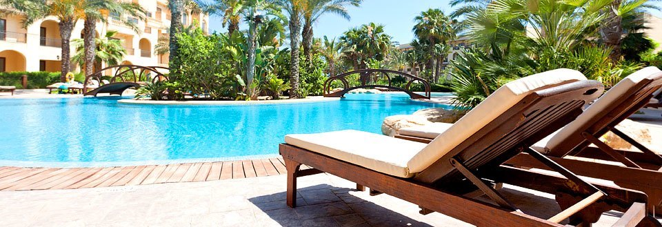 Ett lyxigt semesteranläggning med en pool omgiven av palmer och solstolar under en solig himmel.