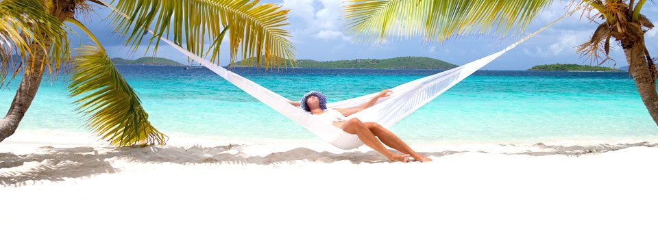 En person vilar i en hängmatta mellan palmer på en pittoresk strand med turkosblått hav.