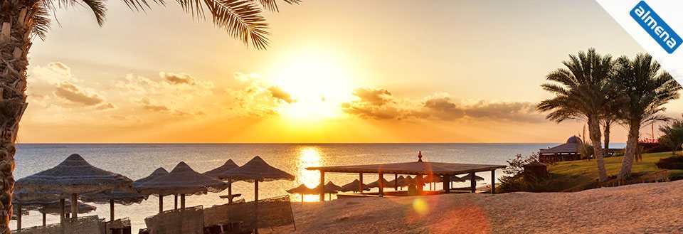 Solnedgång på en strand med palmer och halm parasoller. Havet speglar solens varma färger.