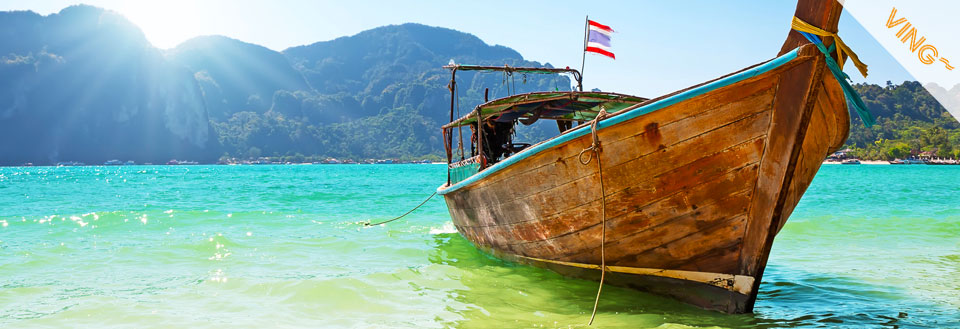 En traditionell träbåt förtöjd vid en tropisk strand med klart blågrönt vatten och berg i bakgrunden.