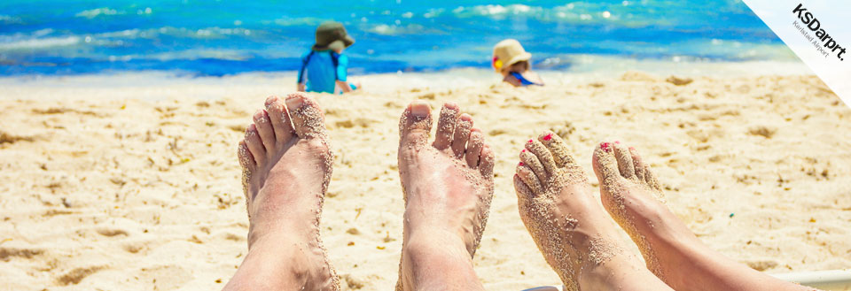 Fyra par fötter täckta med sand på en solig strand med klart blått hav i bakgrunden.