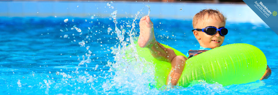 Ett leende barn i solglasögon flyter på en uppblåsbar grön flotte i en pool, vatten stänker runt.