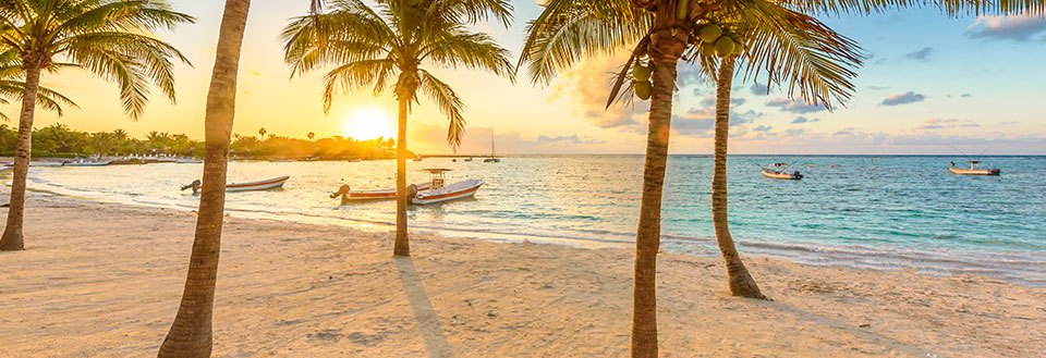 Solnedgång på en tropisk strand med palmer och små båtar i havet.
