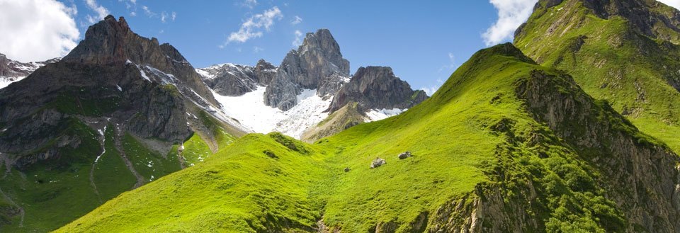 Bilden visar gröna bergssluttningar under en blå himmel med snöklädda bergstoppar i bakgrunden.