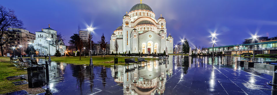 Bilden visar en upplyst kyrka på kvällen med vått trottoar som speglar byggnaderna och ljusen.