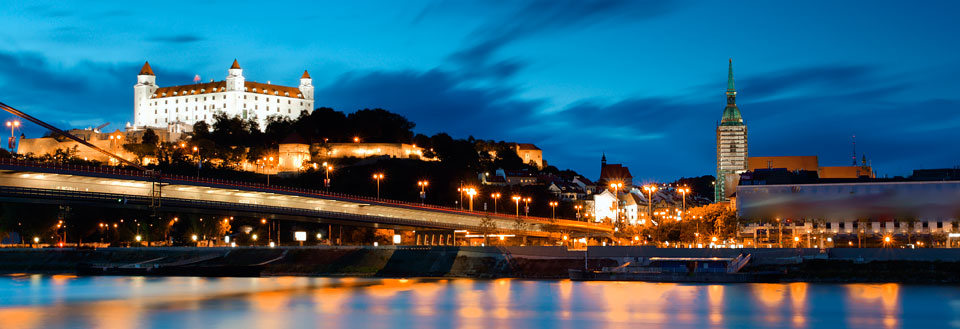 En kvällsbild av Donau i Bratislava med en bro i förgrunden och ett upplyst slott på en kulle i bakgrunden.