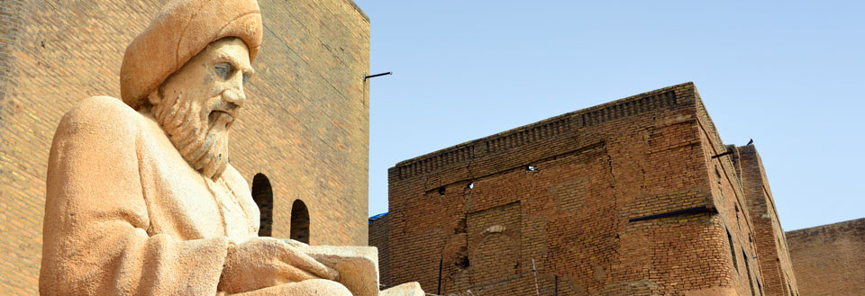 Bilden visar en sandstensskulptur av en man med skägg och turban framför en tegelbyggnad under en klarblå himmel.