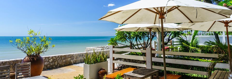 Terrass med havsutsikt: parasoller, stolar och gröna växter framför det klarblå havet.