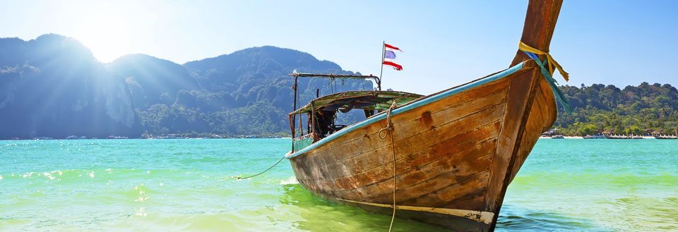 En traditionell träbåt flyter på det klara turkosa vattnet med en bergskedja i bakgrunden under en blå himmel.