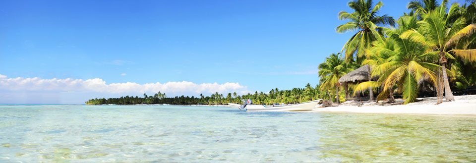 Tropisk strand med palmer, kristallklart vatten och en liten båt. Blå himmel med några moln.