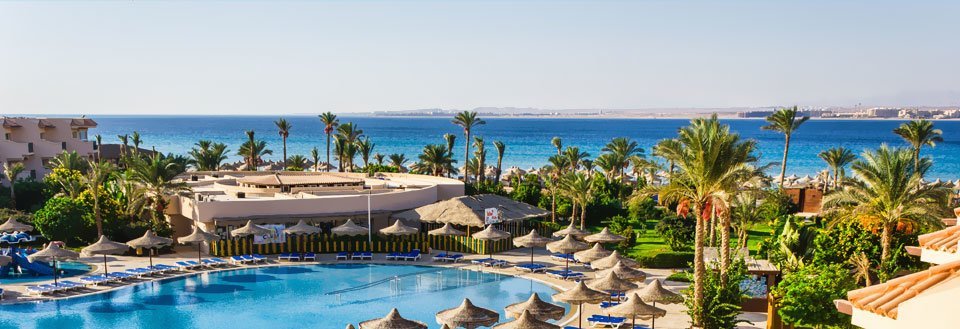 Ett semesteranläggning med pool, palmer och en strandvy i bakgrunden under en klar himmel.