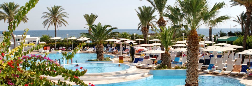 Lyxig semesteranläggning med pooler, palmer och parasoller vid en solig strandkant.