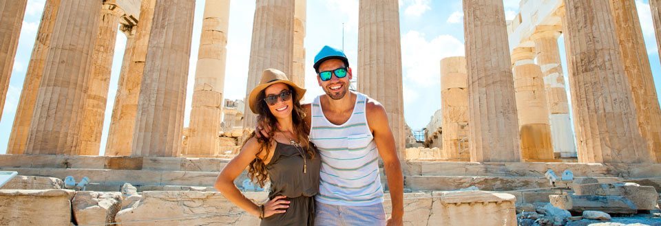 Två leende individer framför antika kolonner under en strålande sol i Athen.
