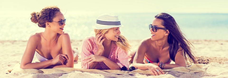 Tre vänner ler och samtalar på en solig strand med klarblå himmel och havet i bakgrunden.