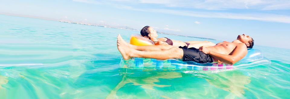 Två personer vilar på luftmadrasser i det kristallklara havet under blå himmel.