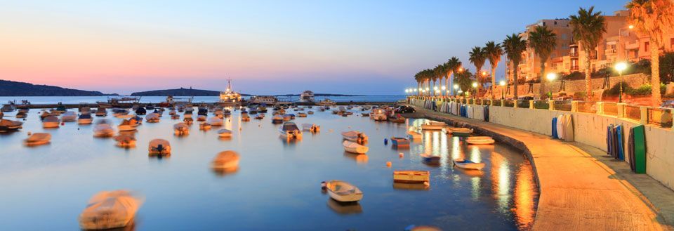 Solnedgång över en hamn i Malta med många små båtar. Vattnet reflekterar den varma himlen och kustlinjen är upplyst.