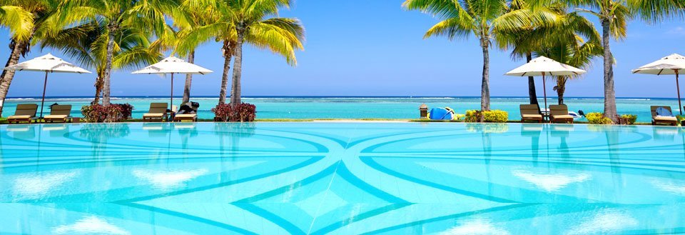 En simbassäng som vetter mot havet, inramad av palmer och solstolar under parasoll.