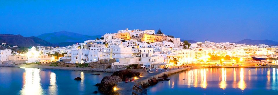 En pittoresk kuststad på Naxos i skymningen med upplysta vita byggnader och reflekterande vatten.