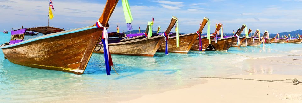 Traditionella Phuket träbåtar med färgglada band förtöjda på en tropisk strand med kristallklart vatten.