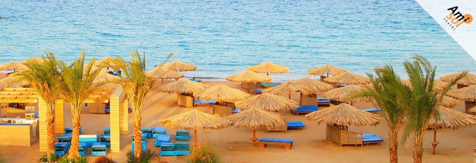 Strand med stråsolparasoller och blå solstolar längs en lugn havskant. Palmer och en bar i bakgrunden.