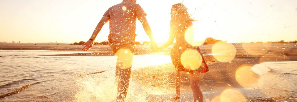 Två personer håller handen och springer längs med strandkanten i solnedgången.