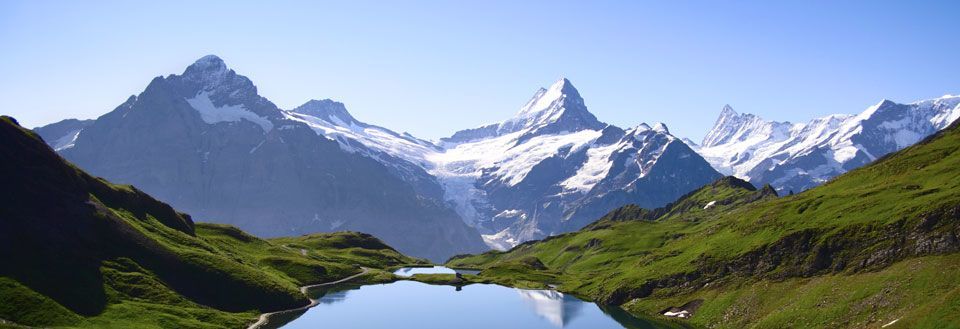Idyllisk bergslandskap med en klarblå sjö i förgrunden och snötäckta toppar i bakgrunden.