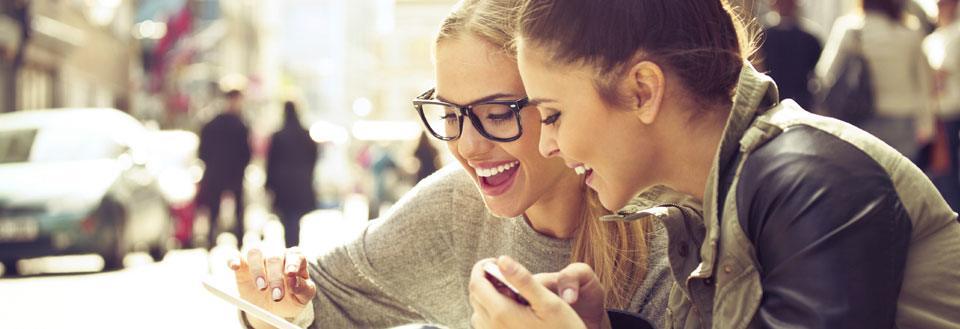 Två leende kvinnor tittar på en smartphone tillsammans på en livlig stadsgata.