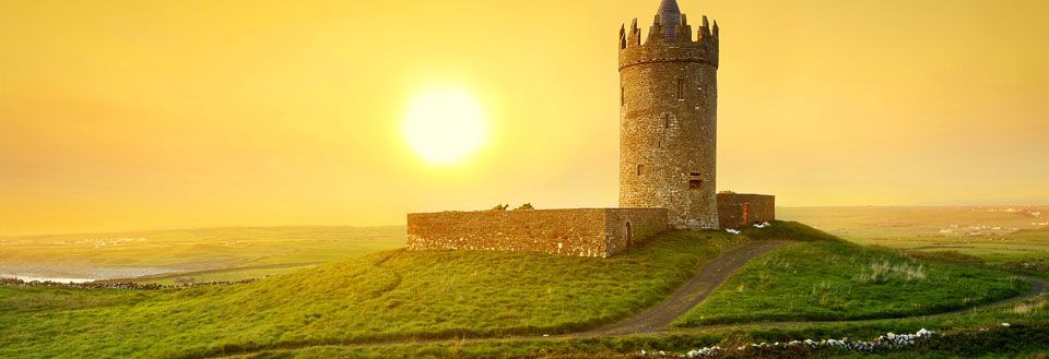 Gammalt stenmur inramar ett medeltida torn på en grön kulle vid solnedgång.