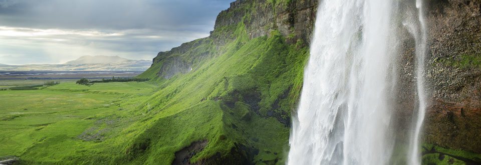 Kraftfullt vattenfall längs en grönbeklädd klippa med utsikt över en bördig slätt.