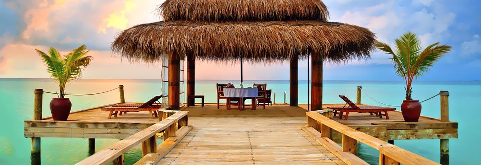 En träbrygga leder ut till en täckt lounge med tak av strå vid ett turkost hav under en färgrik himmel.