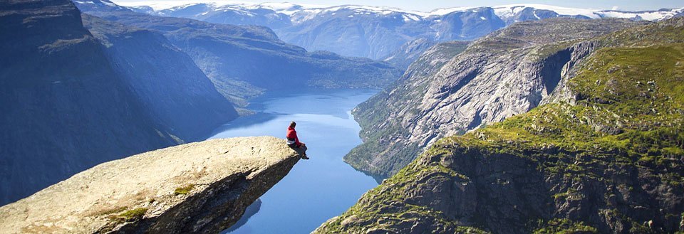En person sitter på kanten av ett bergutsprång med utsikt över en fjord och berg.
