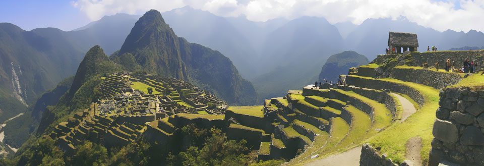 Panoramavy över Machu Picchu med gröna terrasser och dimhöljda berg i bakgrunden.