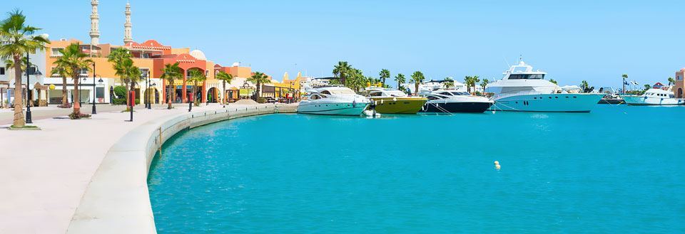 Bild av en solig hamnpromenad i Hurghada  med lyxyachter förtöjda längs kajen framför färgrika byggnader.