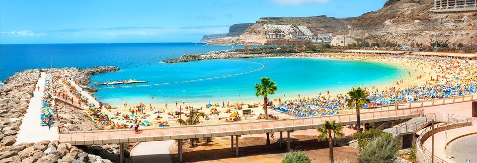 Bilden visar en livlig, solig strand med badgäster, parasoller, och ett klart turkosfärgat hav.