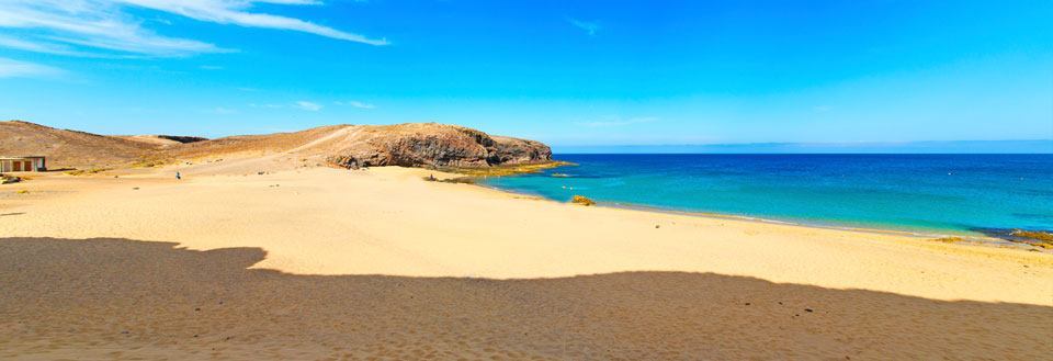 Bild av en vid sandstrand med kristallklart blått vatten och en klippig förgrund under en klar himmel.