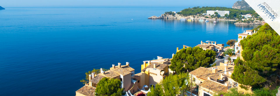 Bilden visar en kustlandskap med terrakottafärgade hus, klarblå himmel och lugnt havsvatten. Bilden visar en kustlandskap med terrakottafärgade hus, klarblå himmel och lugnt havsvatten.