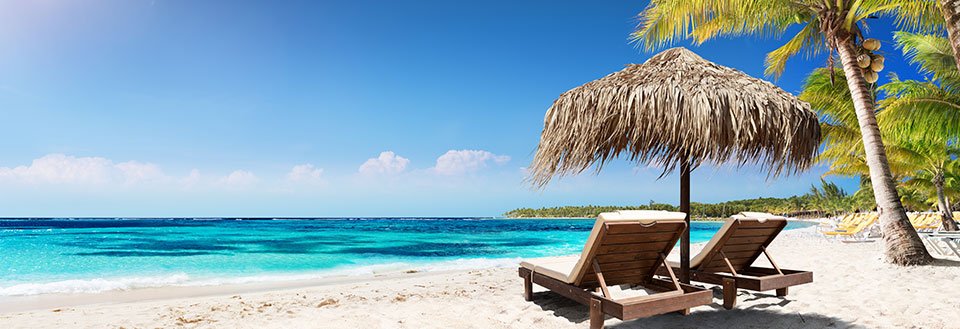 Två solstolar under en stråparasoll på en vit sandstrand med kristallklart blått hav och palmer.