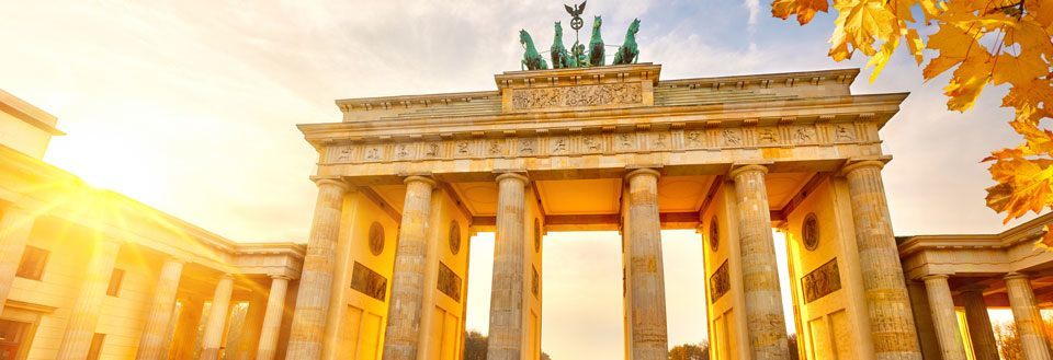 Bilden visar Brandenburger Tor i Berlin badat i det gyllene ljuset från solnedgången, med höstlöv i förgrunden.