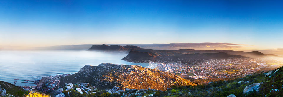 Panoramavy över en kuststad vid solnedgången med berg i bakgrunden och havet i förgrunden.