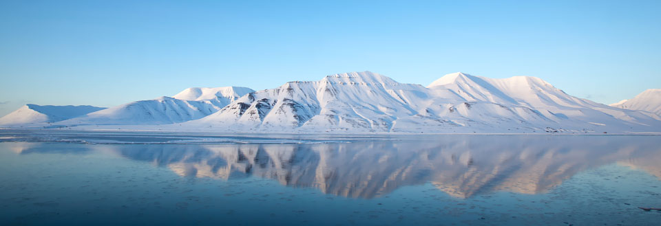 Stilla vattenyta med tydliga speglingar av snötäckta berg och en klarblå himmel.