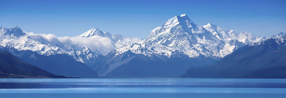 Panoramavy över en spegelblank sjö i Mount Cook nasjonalpark framför majestätiska snötäckta berg under en klarblå himmel.