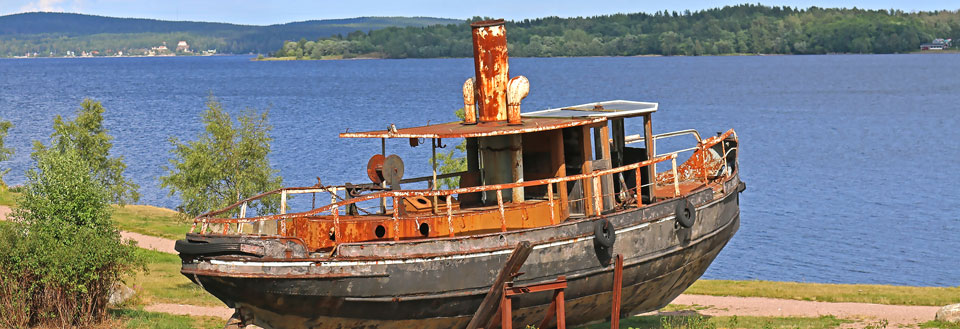 En rostig gammal båt på land med skog och en sjö i bakgrunden.