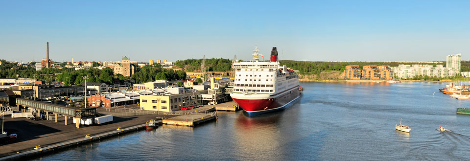 Hamnfront med byggnader, ett stort passagerarfartyg och mindre båtar på en solig dag.